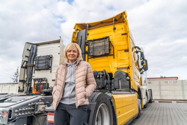 A female truck driver resting against the back wheel of a yellow HGV tractor unit